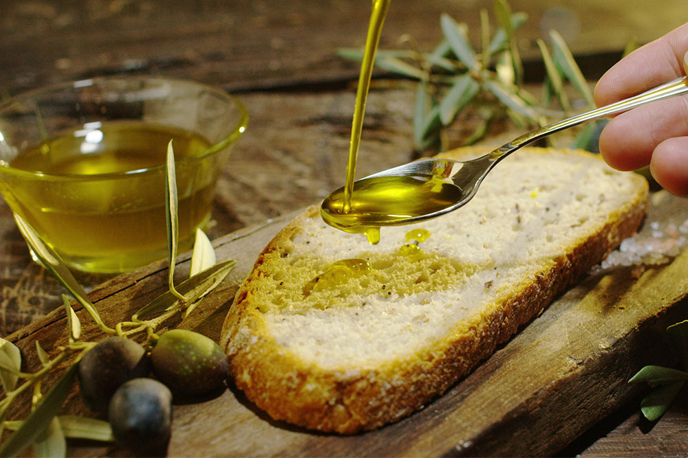 Hand pouring olive oil onto a slice of rustic bread on a wooden board with olives nearby.